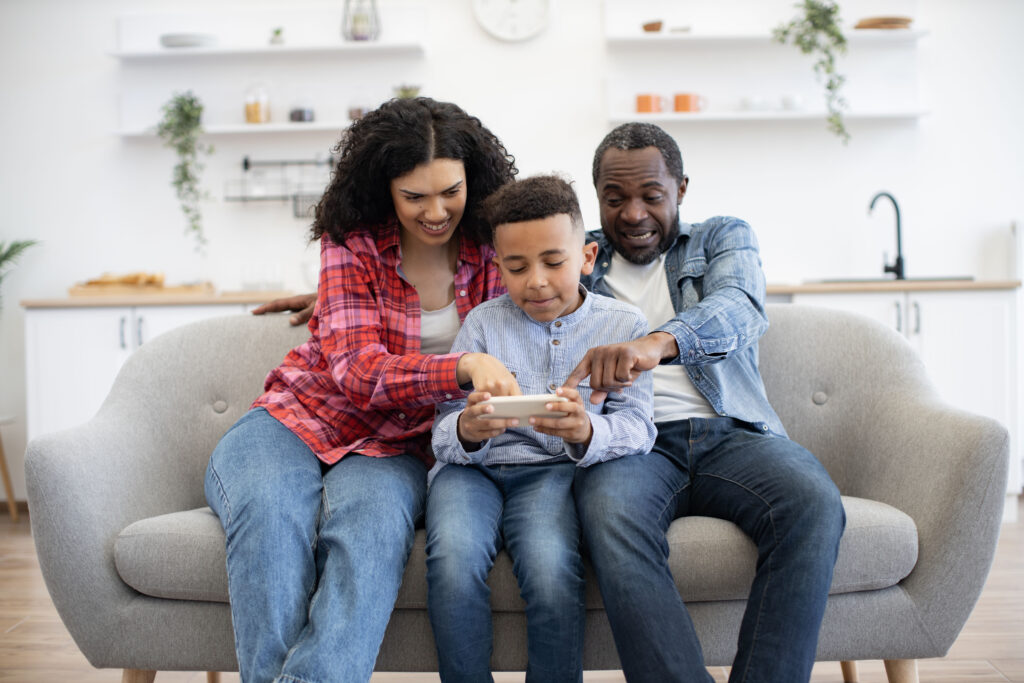 A smiling family sits on a gray couch. The parents, mindful of cell phone boundaries, sit on either side of their young son, who is playing on a tablet. Both parents are looking at the screen and engaging with their son.