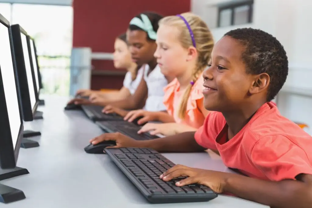 Four children sit in a row at a desk, exploring the best educational sites for kids on desktop computers. They are focused and engaged, with one child in the foreground smiling as they type. The background features a blurred red wall and windows.