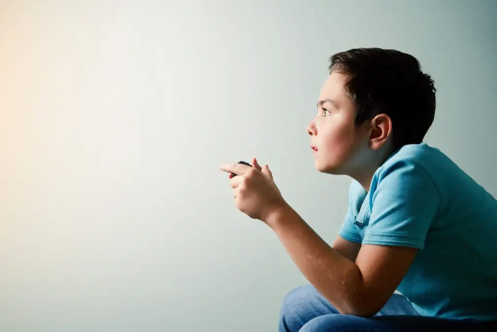 A young boy in a blue shirt sits intently focused, holding a video game controller, seemingly unaware of the dangers of video games. He looks towards a light source on the left, with a neutral background behind him.