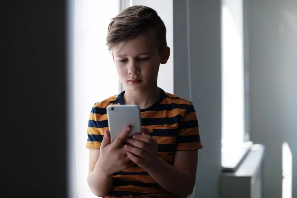 A young boy with short hair and wearing a striped shirt is focused on a smartphone, sitting indoors near a window. The light casts soft shadows, creating a calm atmosphere—one that serves as a gentle reminder that violent media is bad for kids.