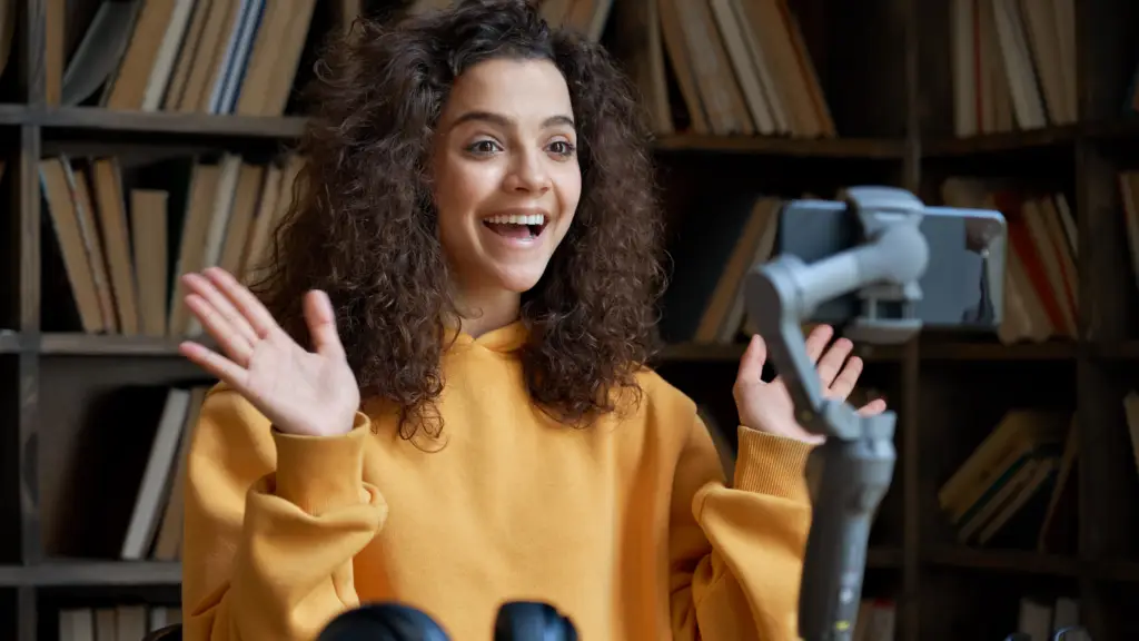 A person with curly hair in a yellow sweater is smiling and gesturing with their hands while looking at a smartphone on a stand, illustrating how smartphones make communication easier, all set against the backdrop of a bookshelf.