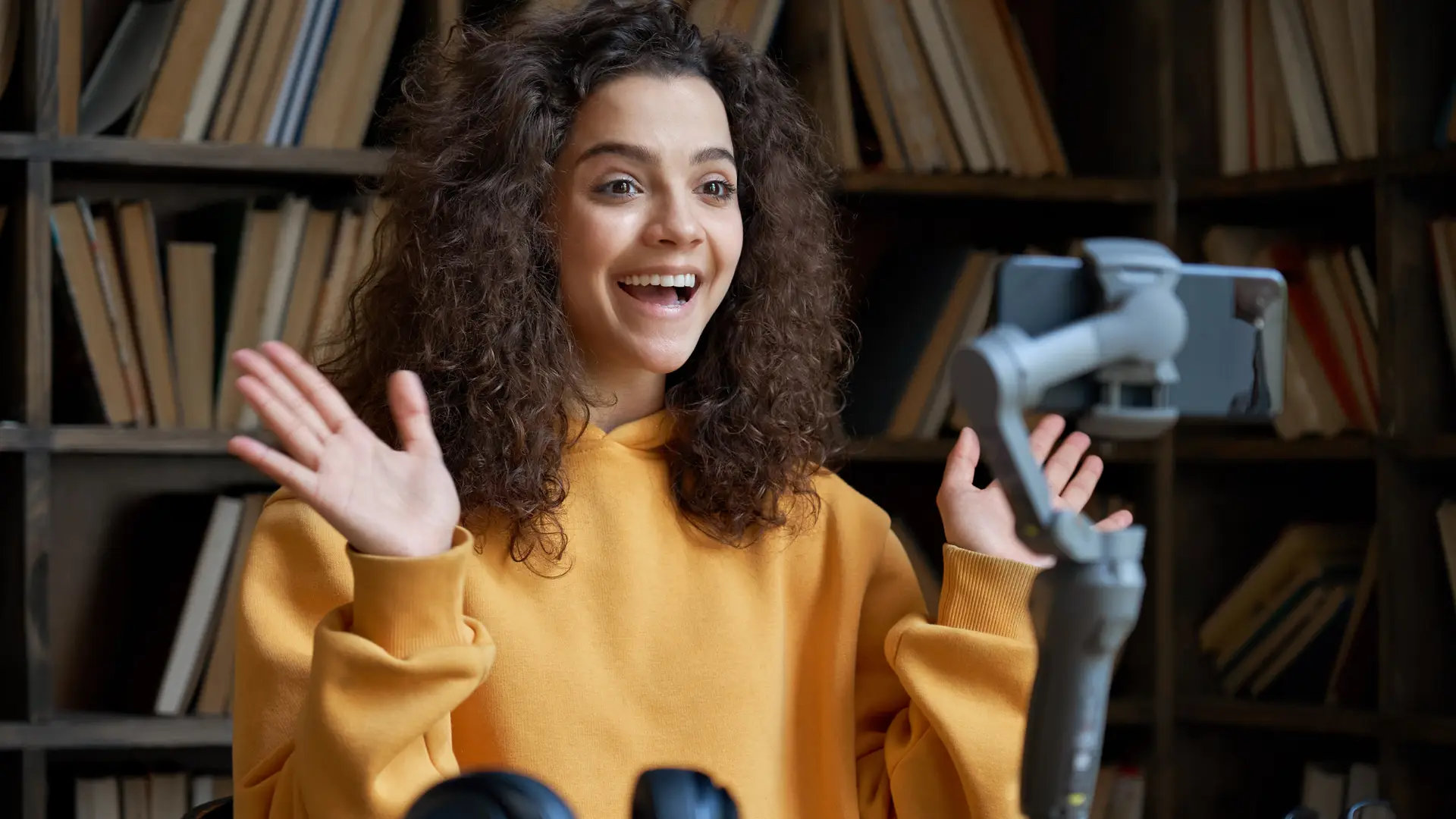 A person with curly hair in a yellow sweater is smiling and gesturing with their hands while looking at a smartphone on a stand, illustrating how smartphones make communication easier, all set against the backdrop of a bookshelf.