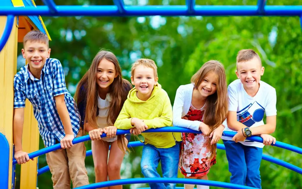 Five cheerful kids are enjoying playtime, leaning over blue bars on a playground structure. They are smiling and wearing casual, colorful clothing. The playground is surrounded by lush green trees, creating a bright and joyful outdoor scene.