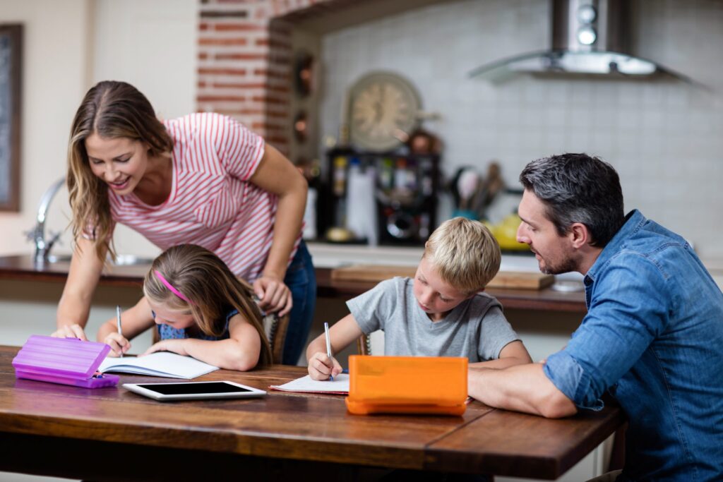 A family sits at a kitchen table, where parental influence is evident as a woman and a man help their children with homework. The kids are writing in notebooks, surrounded by a tablet and pencil cases. The setting is warm and domestic.