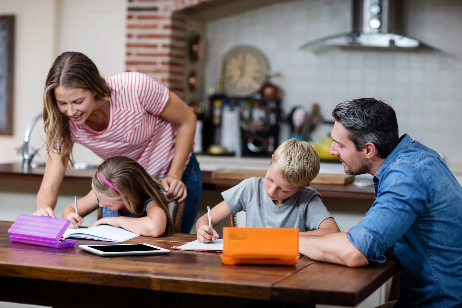 A family sits at a kitchen table, where parental influence is evident as a woman and a man help their children with homework. The kids are writing in notebooks, surrounded by a tablet and pencil cases. The setting is warm and domestic.