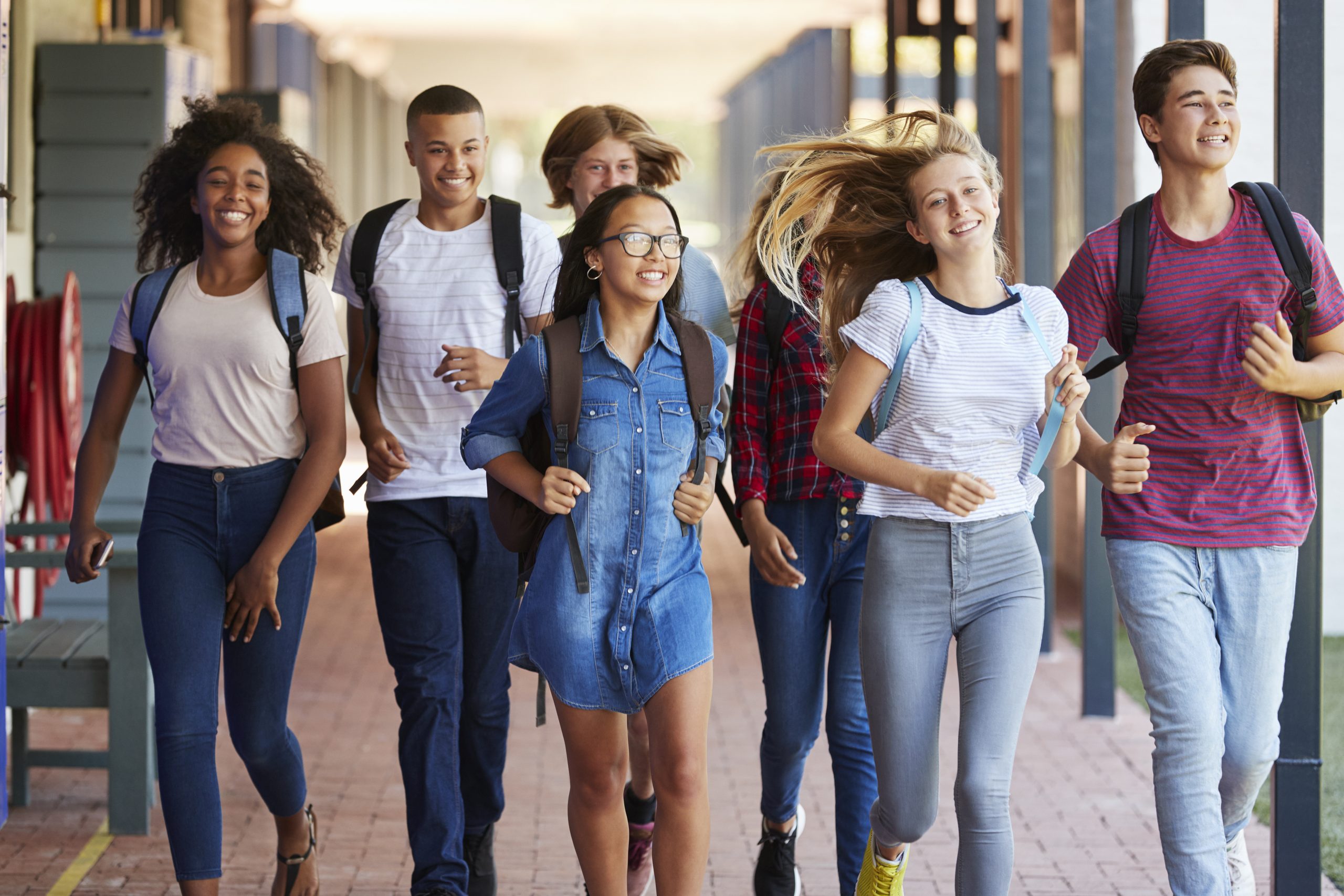 A group of six diverse teenagers, four girls and two boys, walk happily down a covered sidewalk in a polarized society, each carrying backpacks. They look cheerful and energetic, suggesting a school setting or a casual outing.