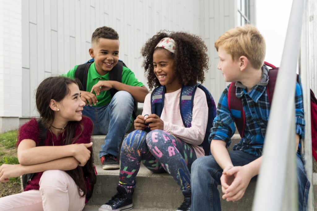 A group of four children sit on outdoor steps, smiling and talking about screen time rules. They appear to be school-aged, with backpacks and casual clothing. The setting suggests a friendly, relaxed atmosphere.