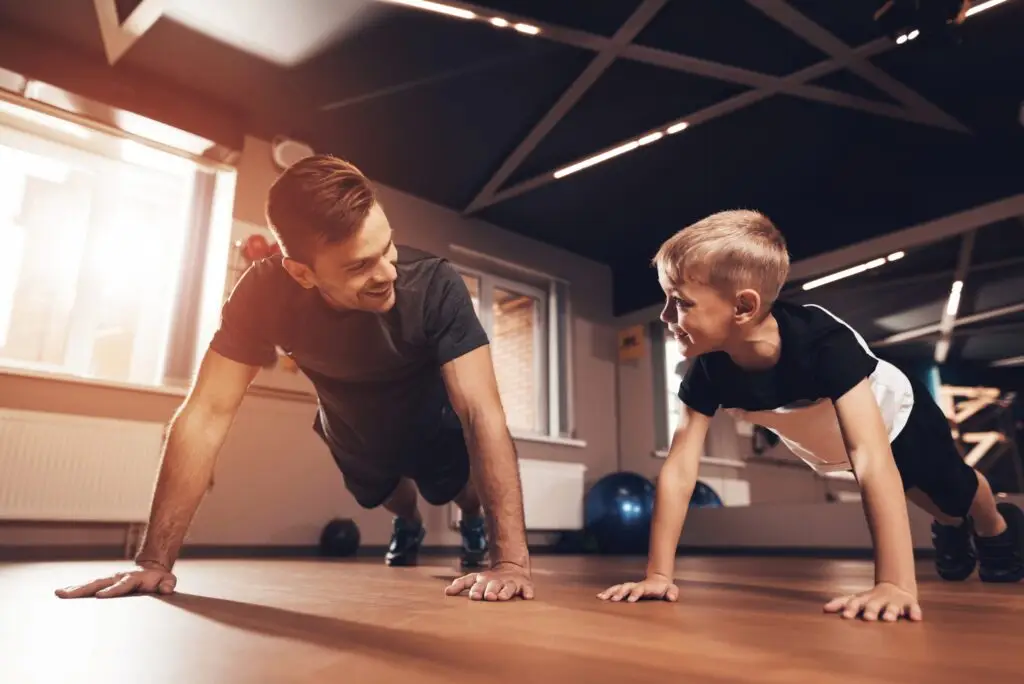 A man and a child are doing push-ups side by side on the wooden gym floor, their smiles shining as brightly as the sun streaming through the windows. Amidst the fitness equipment in the background, they embrace healthy habits, fostering positive self-perception together.