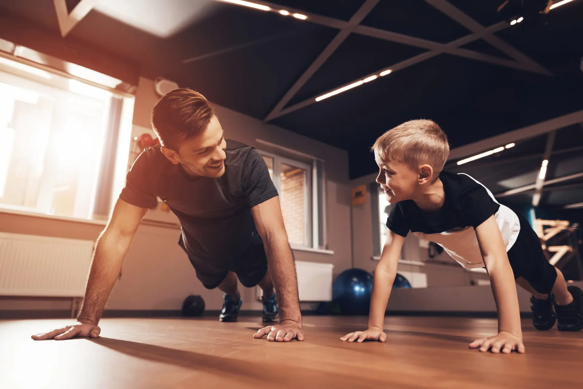 A man and a child are doing push-ups side by side on the wooden gym floor, their smiles shining as brightly as the sun streaming through the windows. Amidst the fitness equipment in the background, they embrace healthy habits, fostering positive self-perception together.