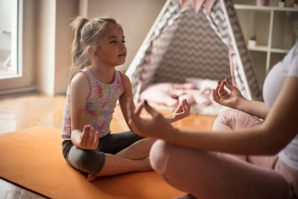 A young girl and an adult sit cross-legged on an orange mat in a cozy room, both practicing meditation with their eyes closed. A patterned play tent is visible in the background—an ideal setting to understand why taking care of your mental health is important.