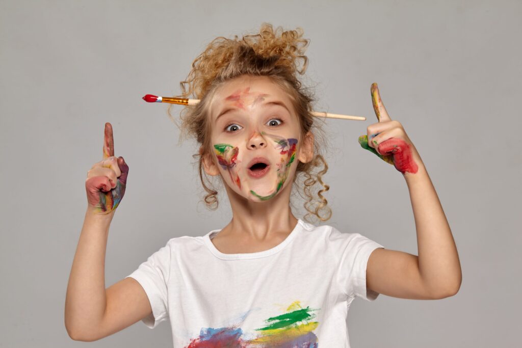 A playful child with curly hair and a paintbrush behind one ear embodies how to cultivate creativity, with colorful paint on their face and hands. Wearing a white shirt splattered in hues, they point both index fingers up, radiating an excited expression.