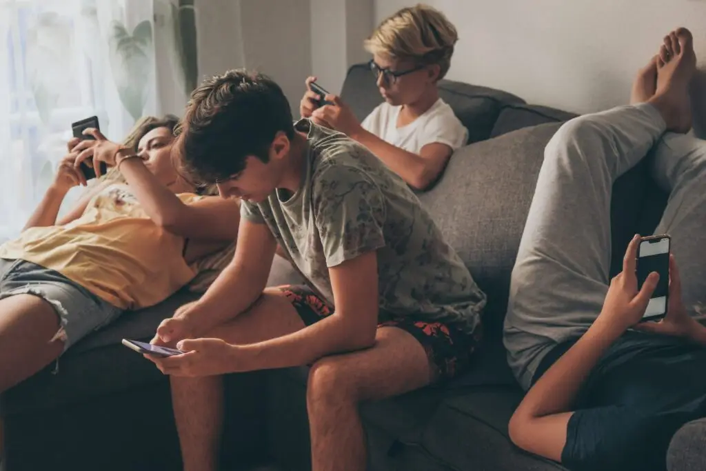 Four teenagers are lounging on a couch, each absorbed in their smartphones, highlighting the quiet loneliness technology can bring. One is lying down with legs up the wall, while others sit casually. The room is softly lit with a window and curtain visible in the background.