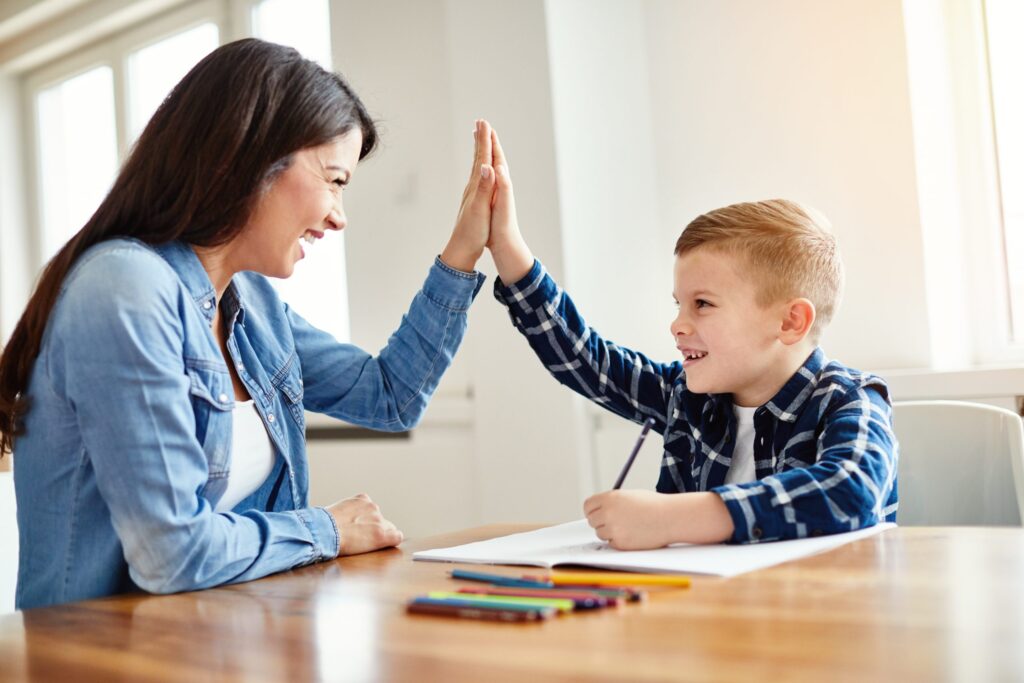 A woman and a young boy sit at a table, smiling and high-fiving as they share a moment of joy. The boy is holding a pencil with a notebook in front of him, colored pencils scattered on the table, as they engage in teaching kids self-discipline through a fun learning activity.