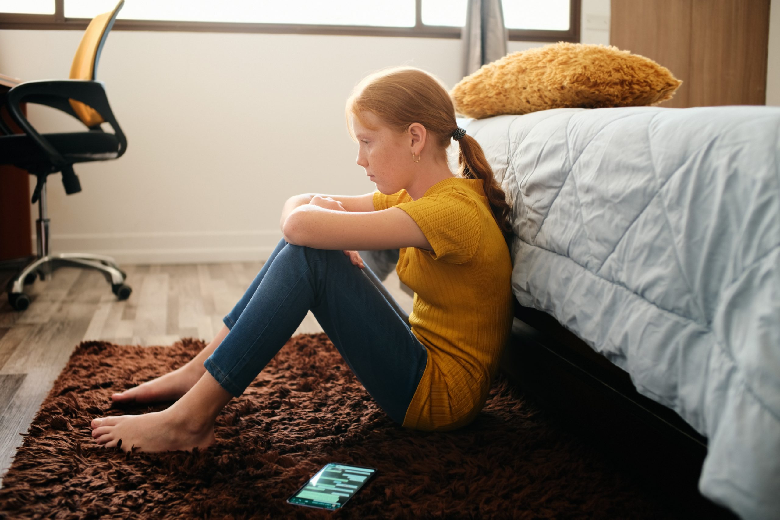 A young girl with red hair sits thoughtfully on a brown carpet in a bedroom, leaning against a bed. Her hands rest on her knees as she contemplates the cyber bullying law. A smartphone lies on the floor nearby, while wooden flooring and a desk chair complete the scene.