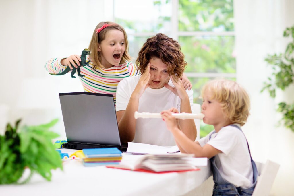 A woman looks stressed while working on a laptop at a table, striving to find balance in life. A young girl energetically plays with a dinosaur toy, while a young boy plays a flute nearby. The scene is set in a bright room with plants and books.