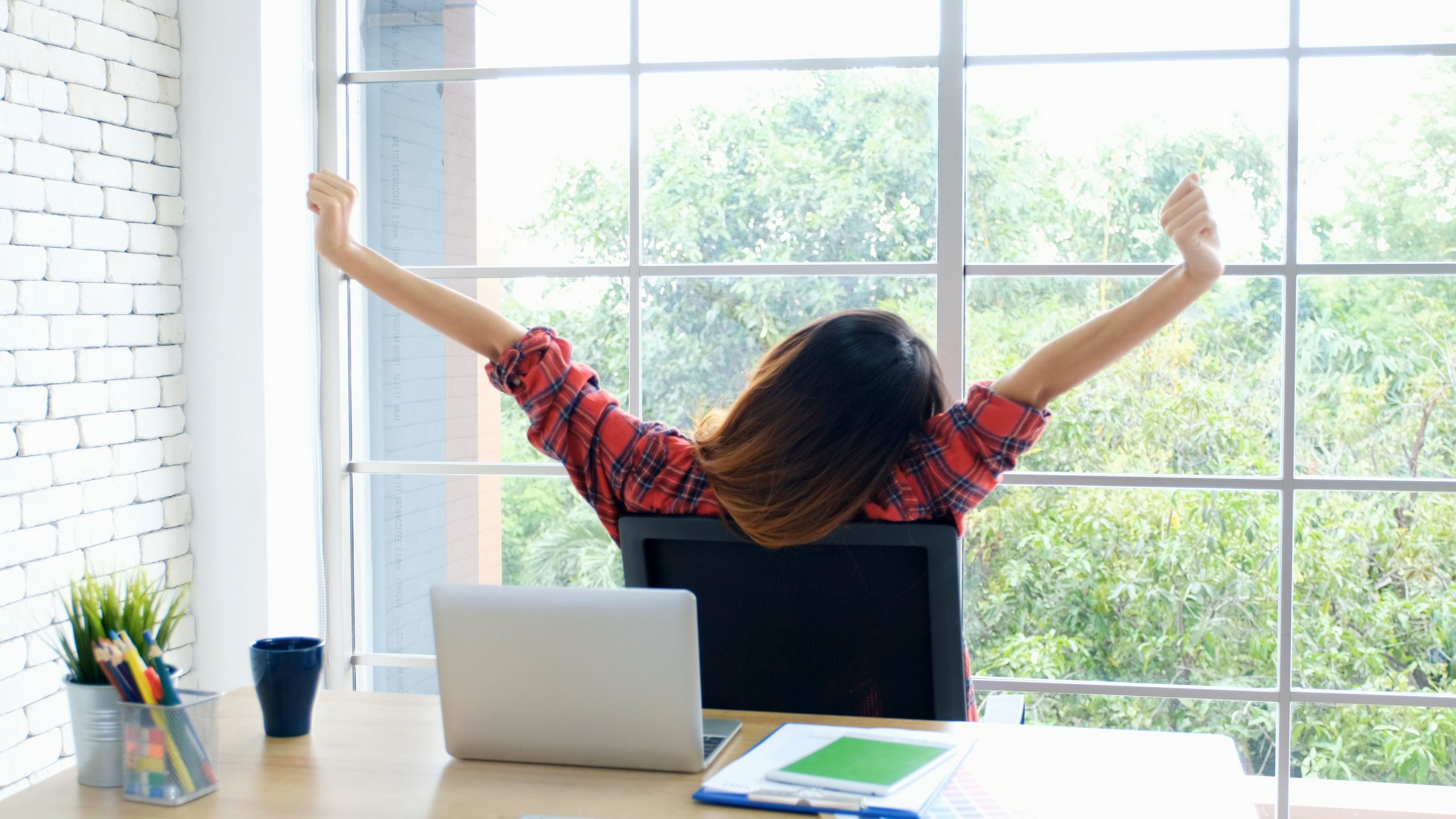 A person wearing a red plaid shirt stretches with arms raised while sitting at a desk, taking a technology break. A laptop, documents, and a potted plant are on the desk. The large window behind shows green trees, suggesting a serene office or home workspace.