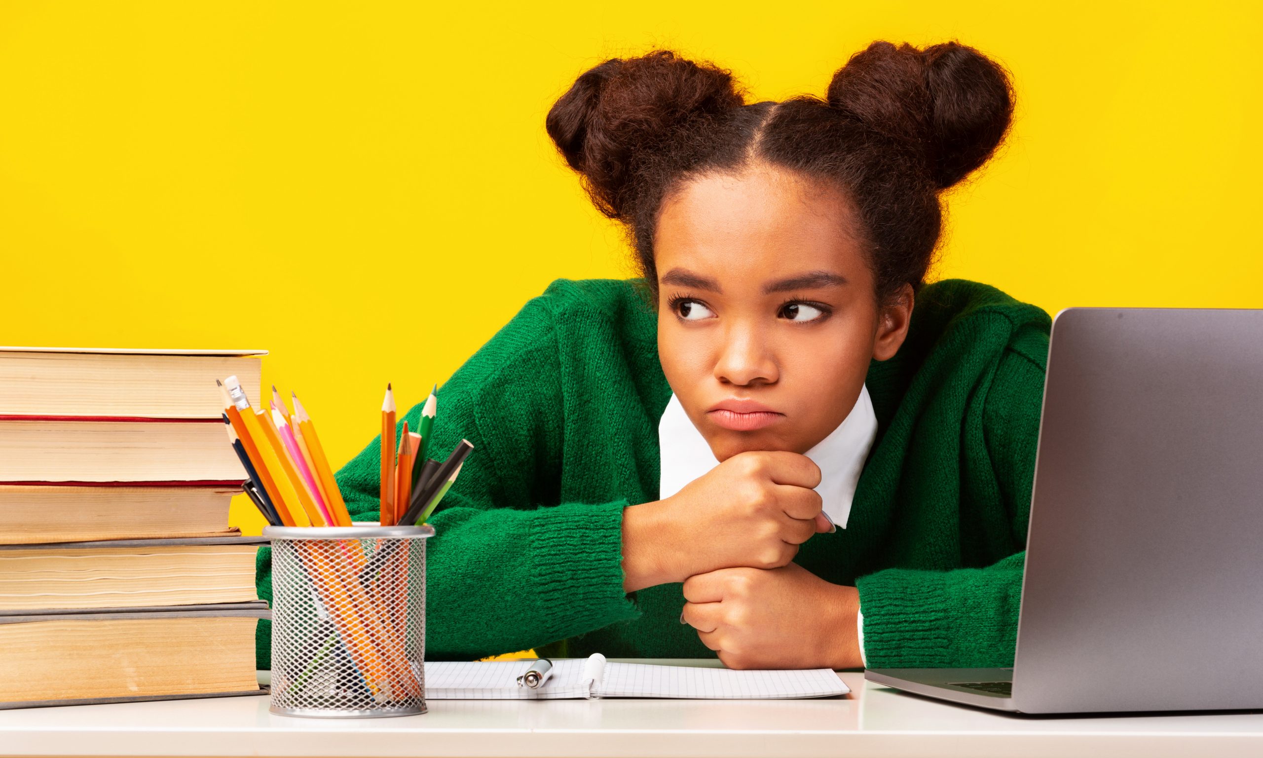 A girl in a green sweater sits at a desk with a stack of books, a pencil holder, and a laptop. She looks contemplative, resting her chin on her fists. It's clear she could use some mental health activities for kids to help ease her frustration against the bright yellow background.