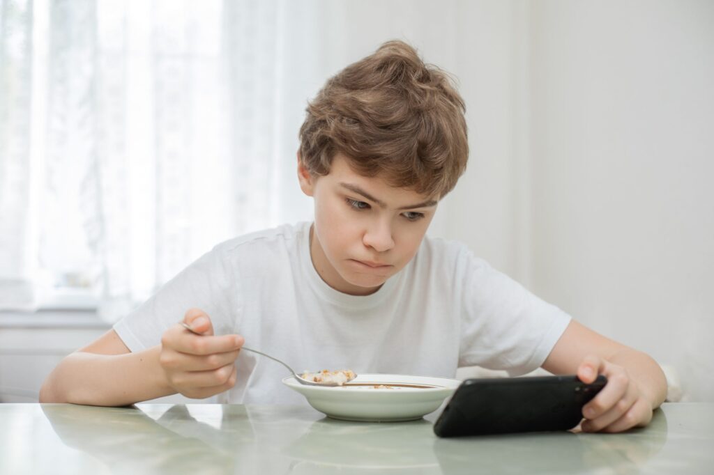 A young boy in a white t-shirt sits at a table, clutching a spoon in one hand and absorbed by his smartphone in the other. As he gazes intently at the screen, it's clear that this quiet moment might be a stepping stone on how to break mobile phone addiction. The soft light filters through sheer curtains.