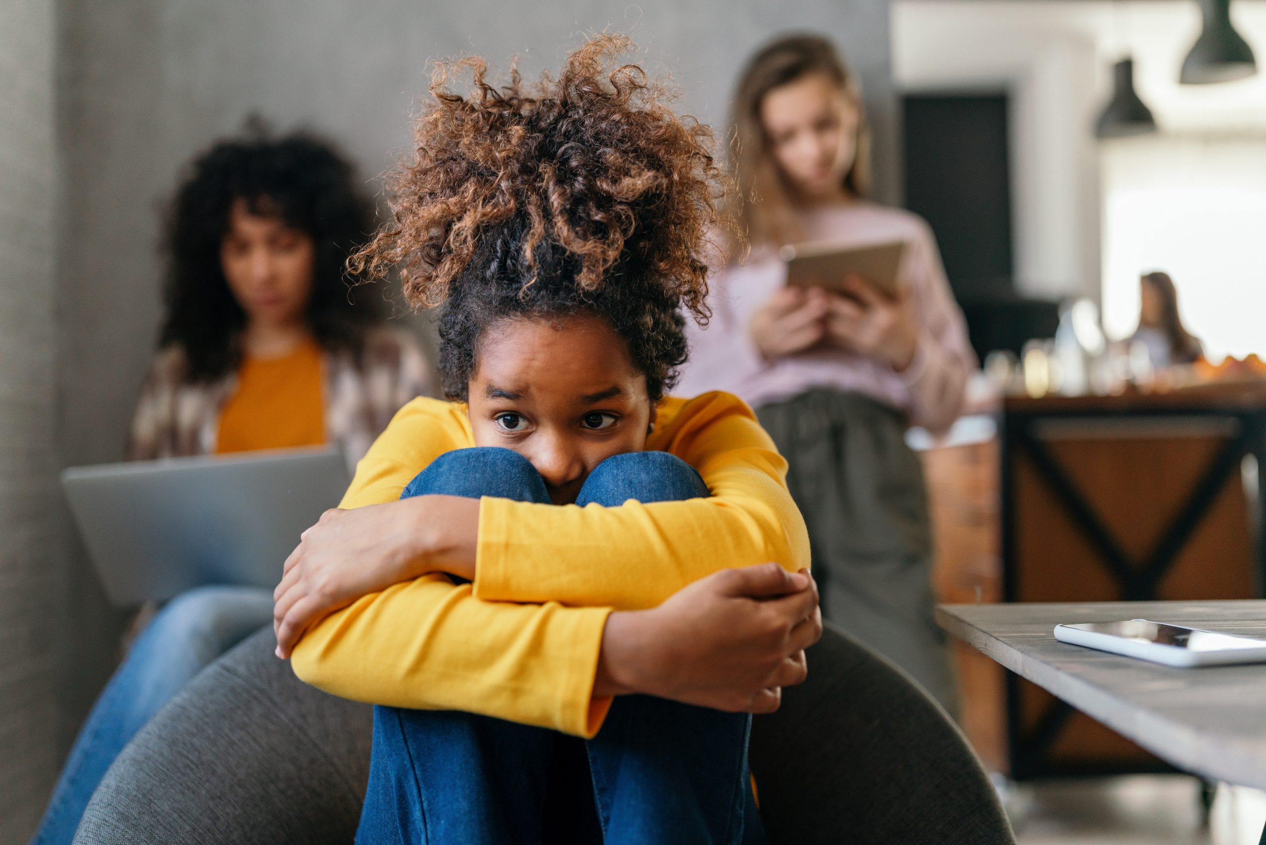 A young girl with curly hair, wearing a yellow sweater, sits with her knees pulled up, looking thoughtful. In the background, amid the subtle hum of technology, two women are absorbed in their devices. The setting appears to be a cozy living room tinged with an undercurrent of anxiety.