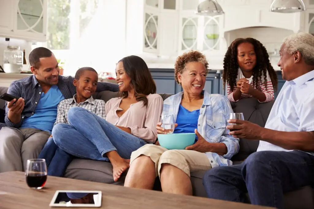 A joyful multigenerational family sits together on a couch, their smiles and conversations highlighting genuine personal interaction. Drinks in hand and snacks in reach, they enjoy each other's company. In the foreground, a tablet and a glass of red wine rest on the table.