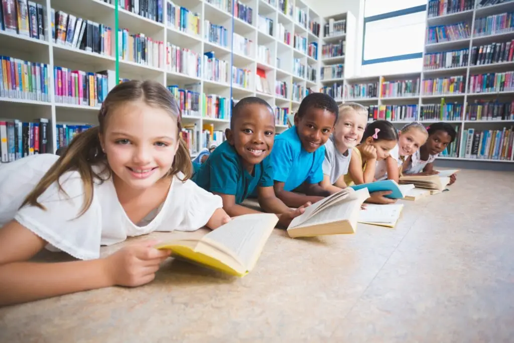 Six children lie on the library floor in a row, smiling and reading books—an ideal scene for learning how to encourage your child to read. The bright lighting and colorful shelves behind them create a cheerful atmosphere, inspiring young minds to explore the joy of reading.