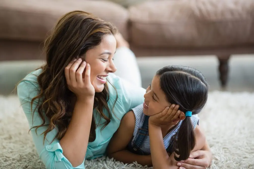 A woman and a young girl lie on a light-colored carpet, facing each other with warm smiles. The woman has long brown hair, while the girl sports dark pigtails. They share a moment that reflects innocence and trust, much like knowing how to protect children online can offer peace of mind.