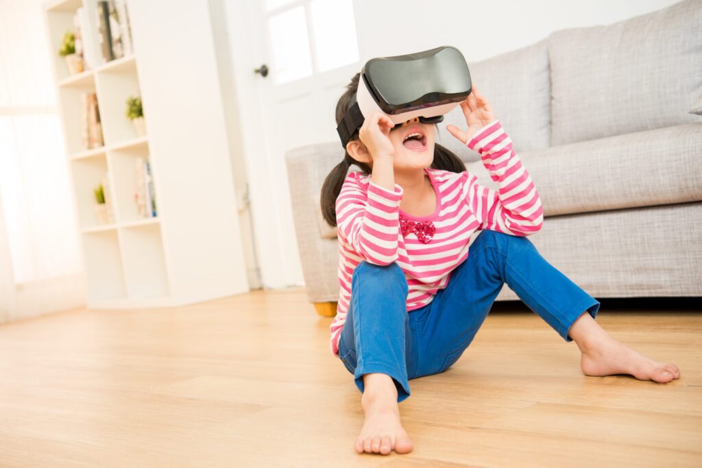 A child wearing a virtual reality headset sits excitedly on the wooden floor of a cozy living room, immersing themselves in digital worlds. They don a pink and white striped shirt with blue pants, against the backdrop of a couch and bookshelf—a scene blending playtime with hints of modern social isolation.