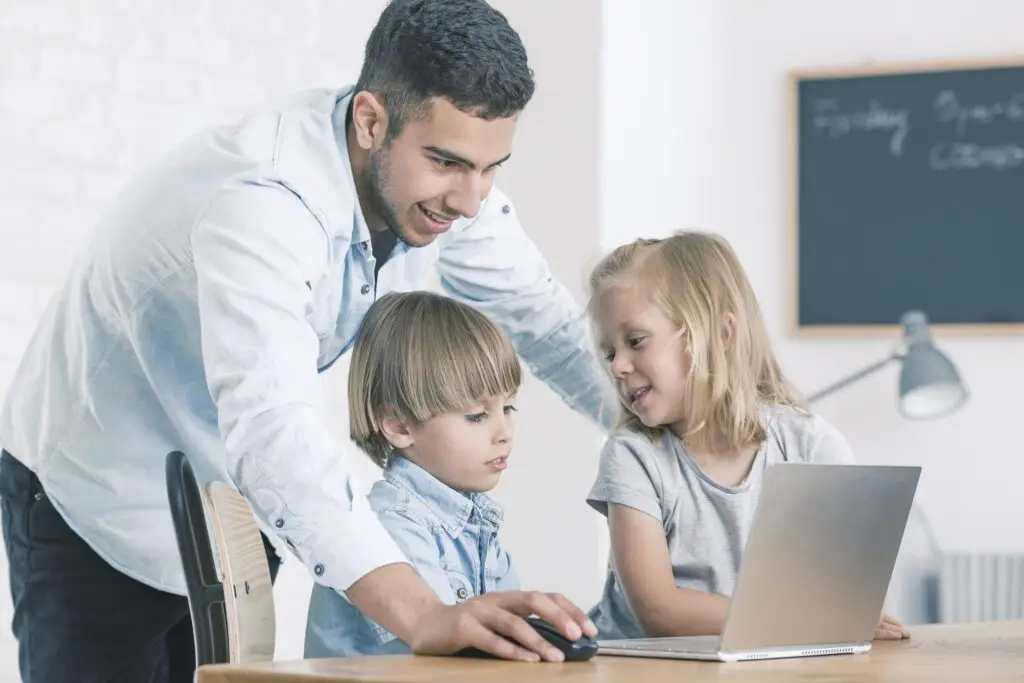 A man and two children are gathered around a laptop. As he demonstrates how to navigate safely online, the boy attentively uses the mouse while the girl looks on with interest. They are in a bright room with a chalkboard in the background, highlighting their focus on digital security.