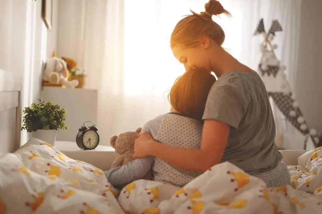 A woman and child sit on a bed in a warmly lit room, embracing with warmth and assurance that they are good enough. The child holds a stuffed bear, as an alarm clock sits on the bedside table. Sunlight filters through the window, highlighting the fox-patterned bedspread in their cozy sanctuary.