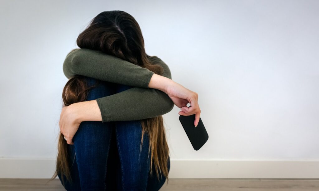 A person sits on the floor with their knees drawn up and arms wrapped around them, lost in social media on a smartphone. They wear a green sweater and jeans, face hidden from view. The backdrop is a plain, light-colored wall as they navigate the digital world in solitude.