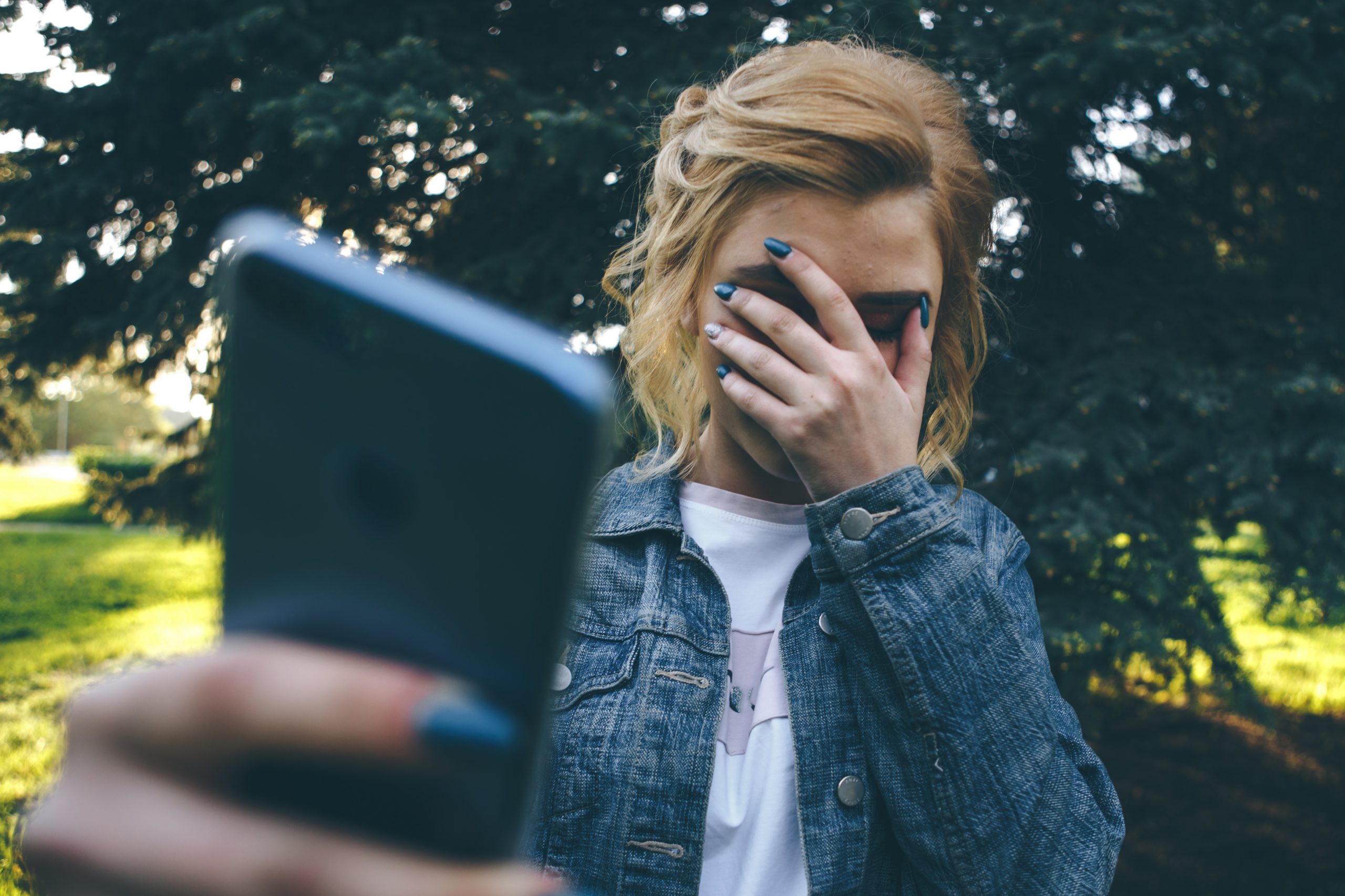 A person with blonde hair in a denim jacket is outdoors taking a selfie, capturing the lush greenery and trees behind. While covering their face with one hand and holding a smartphone with the other, they remind us of the unseen risks, like the dangers of teen sexting.