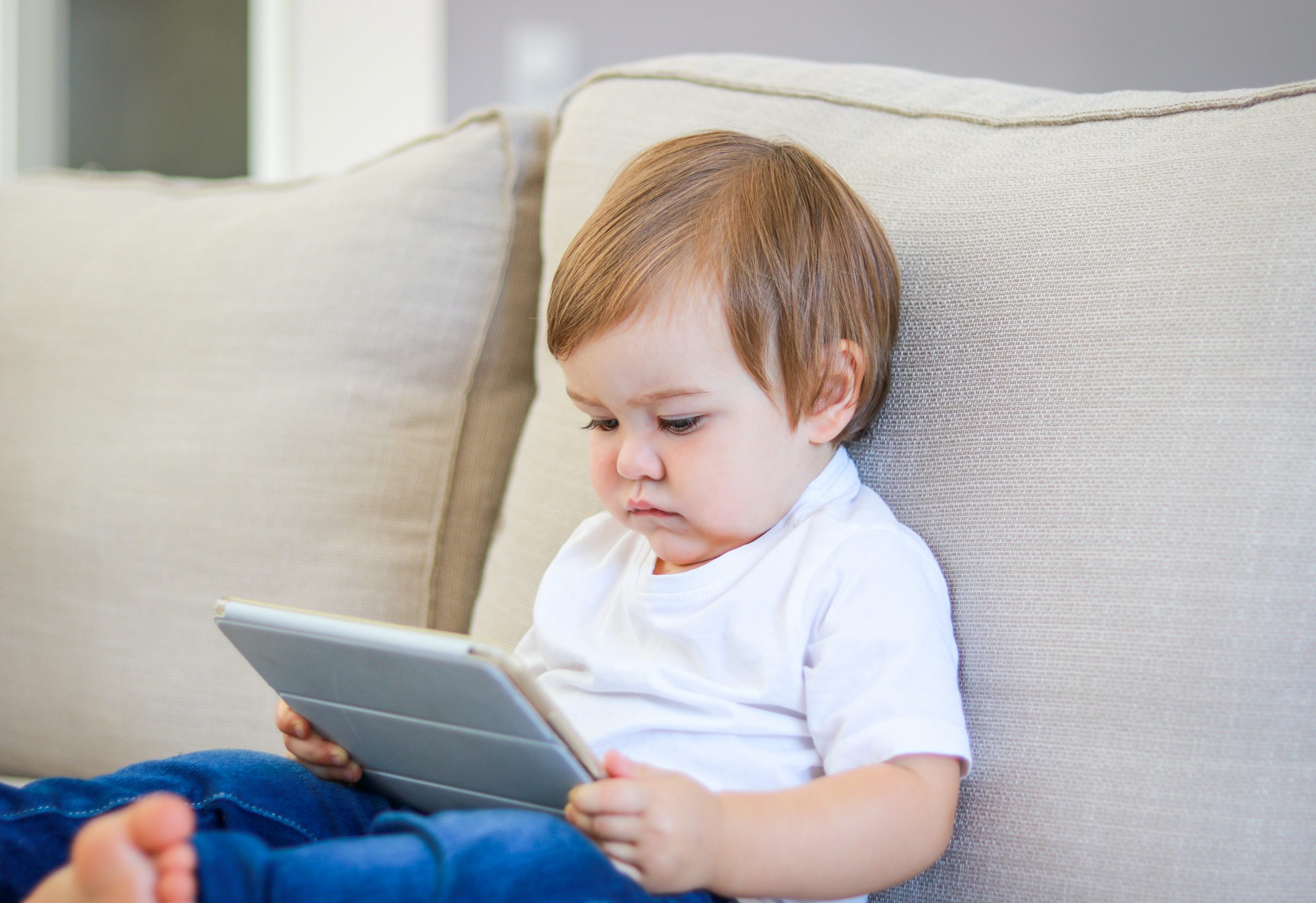 A toddler with brown hair sits on a beige sofa, engrossed in a tablet held in both hands. The child wears a white shirt and blue jeans, with bare feet visible. The softly blurred background underscores the child's focus, prompting thoughts on how young is too young for social media engagement.