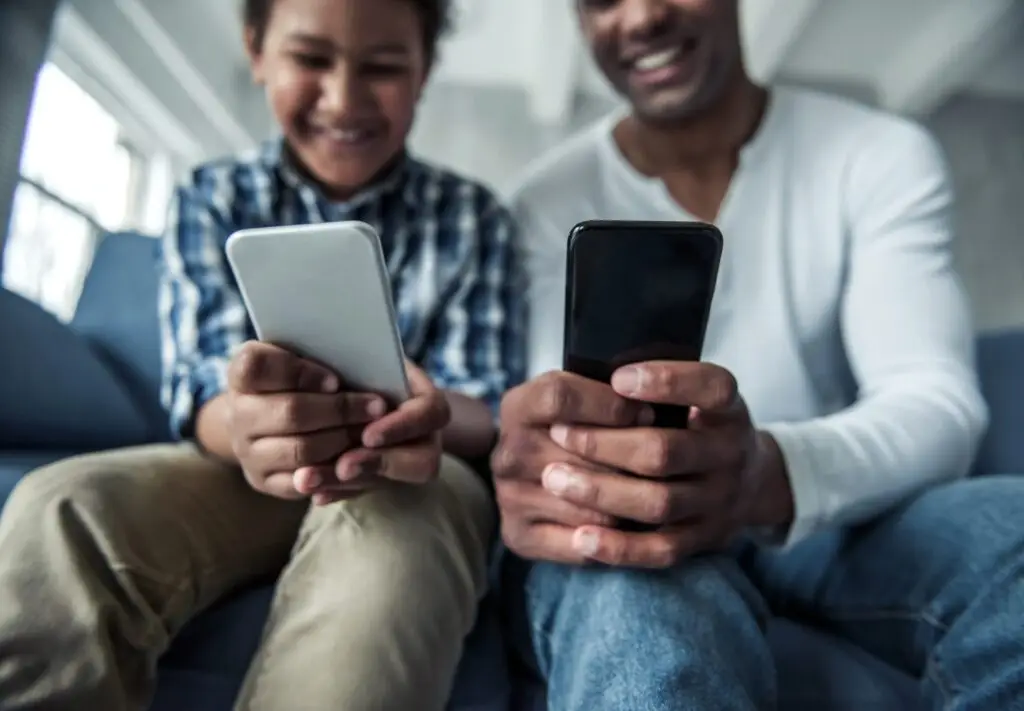 A child and an adult sit on a couch, both smiling and managing their screen time with smartphones. The child wears a plaid shirt and beige pants, while the adult sports a white long-sleeve. They appear to be enjoying their time together in the bright room.