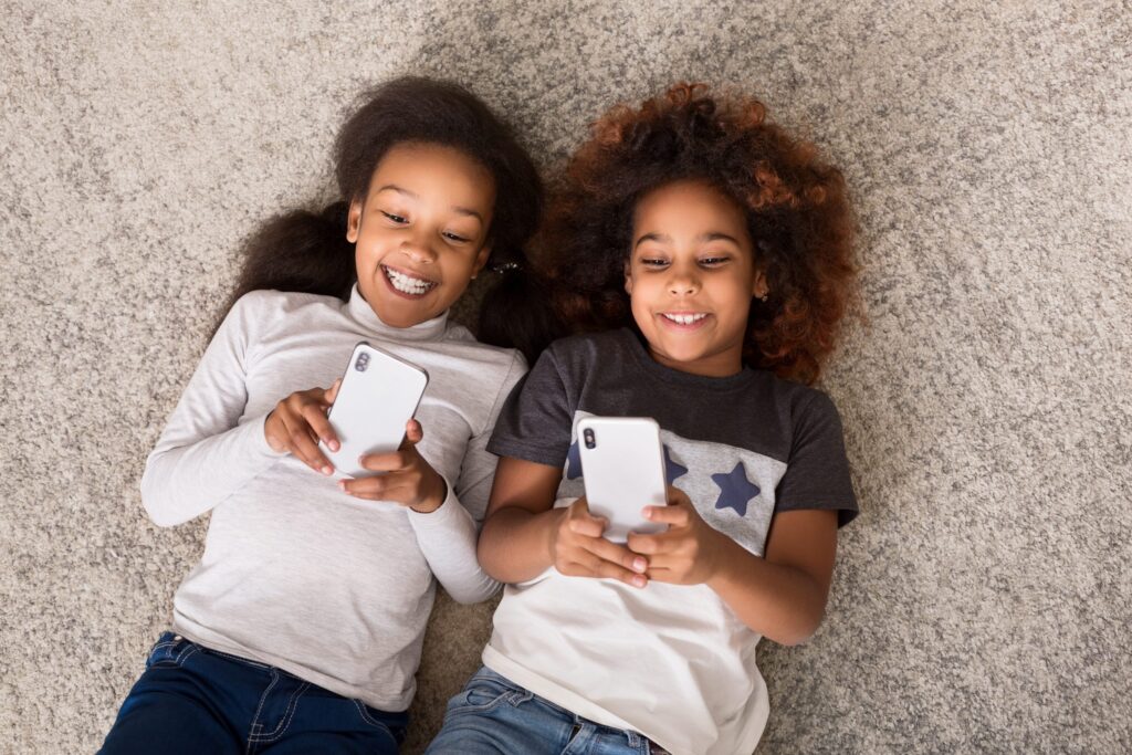 Two kids are lying on a carpet, smiling and using smartphones with safe apps for kids. They appear relaxed and engaged with their devices.