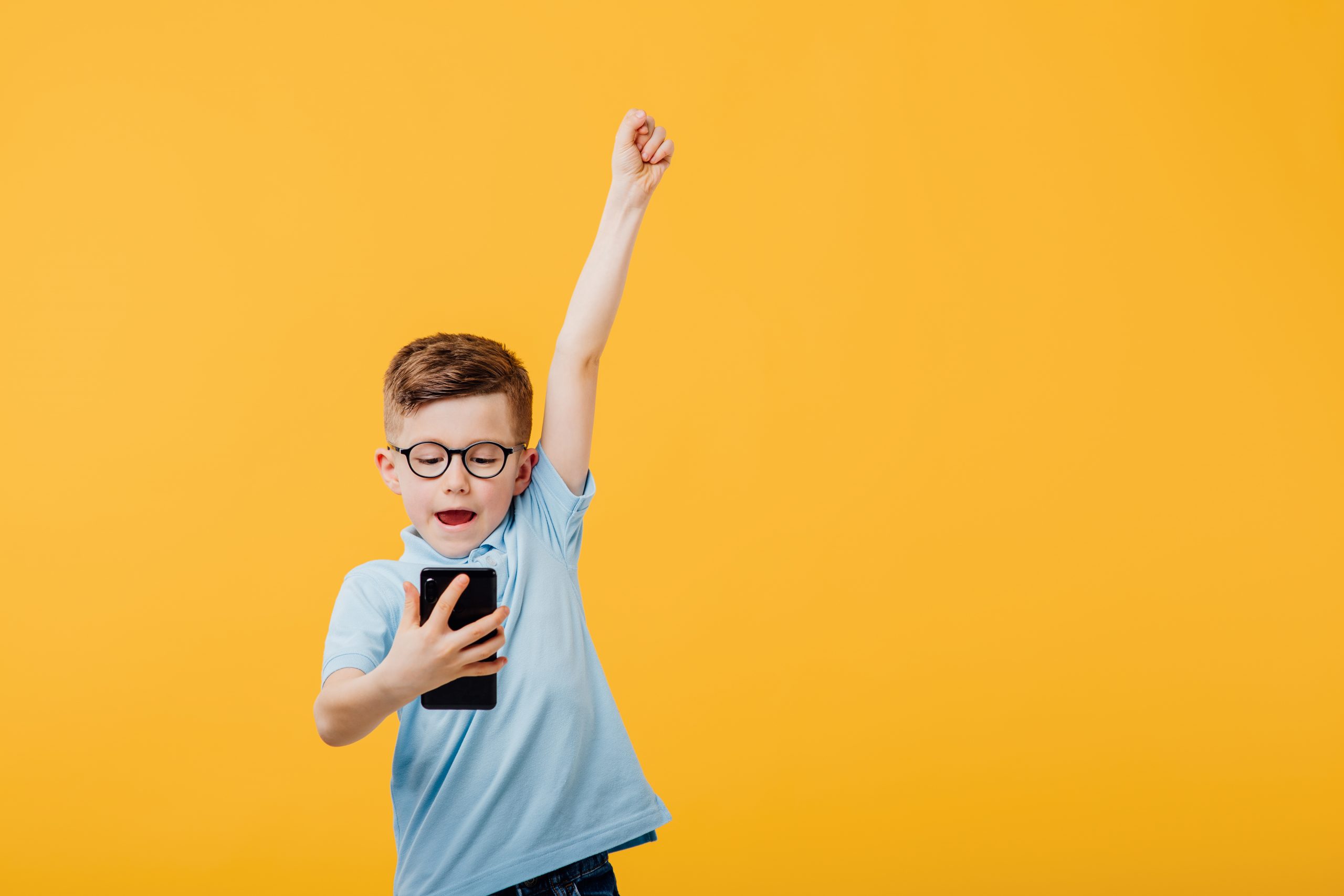 A young boy wearing glasses and a blue shirt excitedly looks at his smartphone, celebrating a victory in maintaining healthy technology habits, with one arm raised against a solid yellow background.