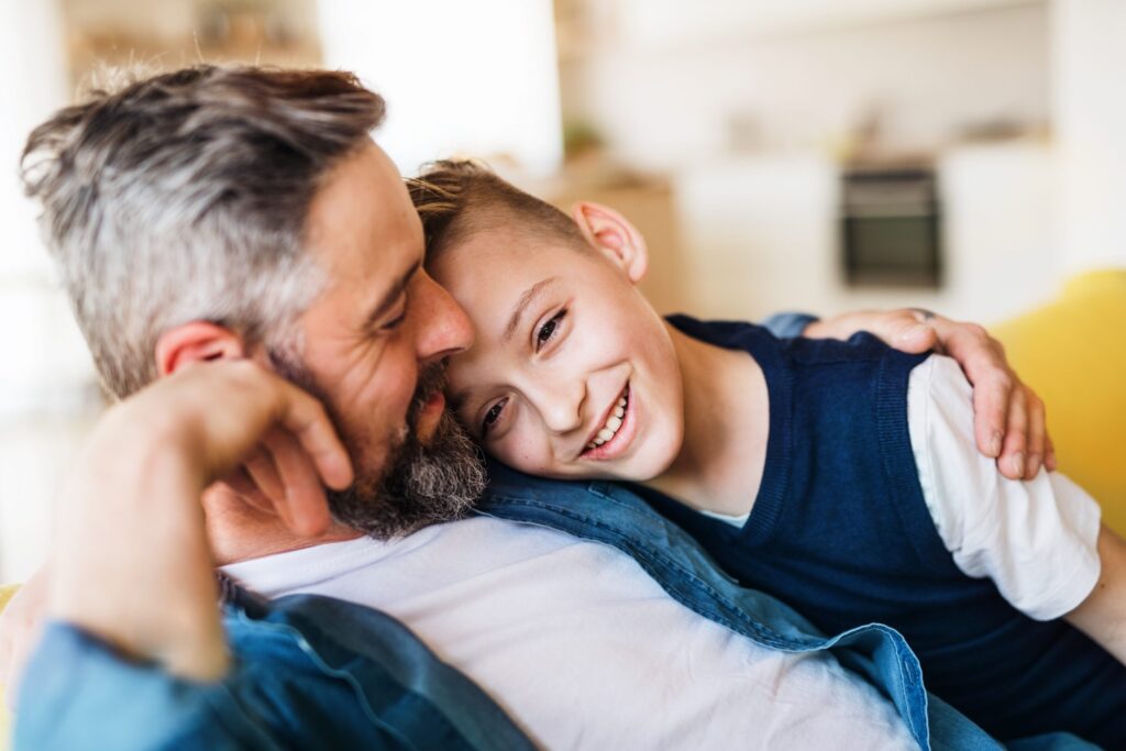 A man and a child are sitting close together on a couch, smiling and embracing, away from screen time. The man has gray hair and a beard, while the child has short blond hair. They appear happy and relaxed in their cozy home setting.