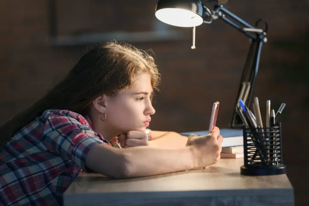 A young person, seemingly absorbed by the smartphone in their hand, sits at a desk lit by a lamp. Clad in a plaid shirt, they lean on their arm amidst pens and a blurred backdrop—an image reflecting how children can become so easily addicted to technology.