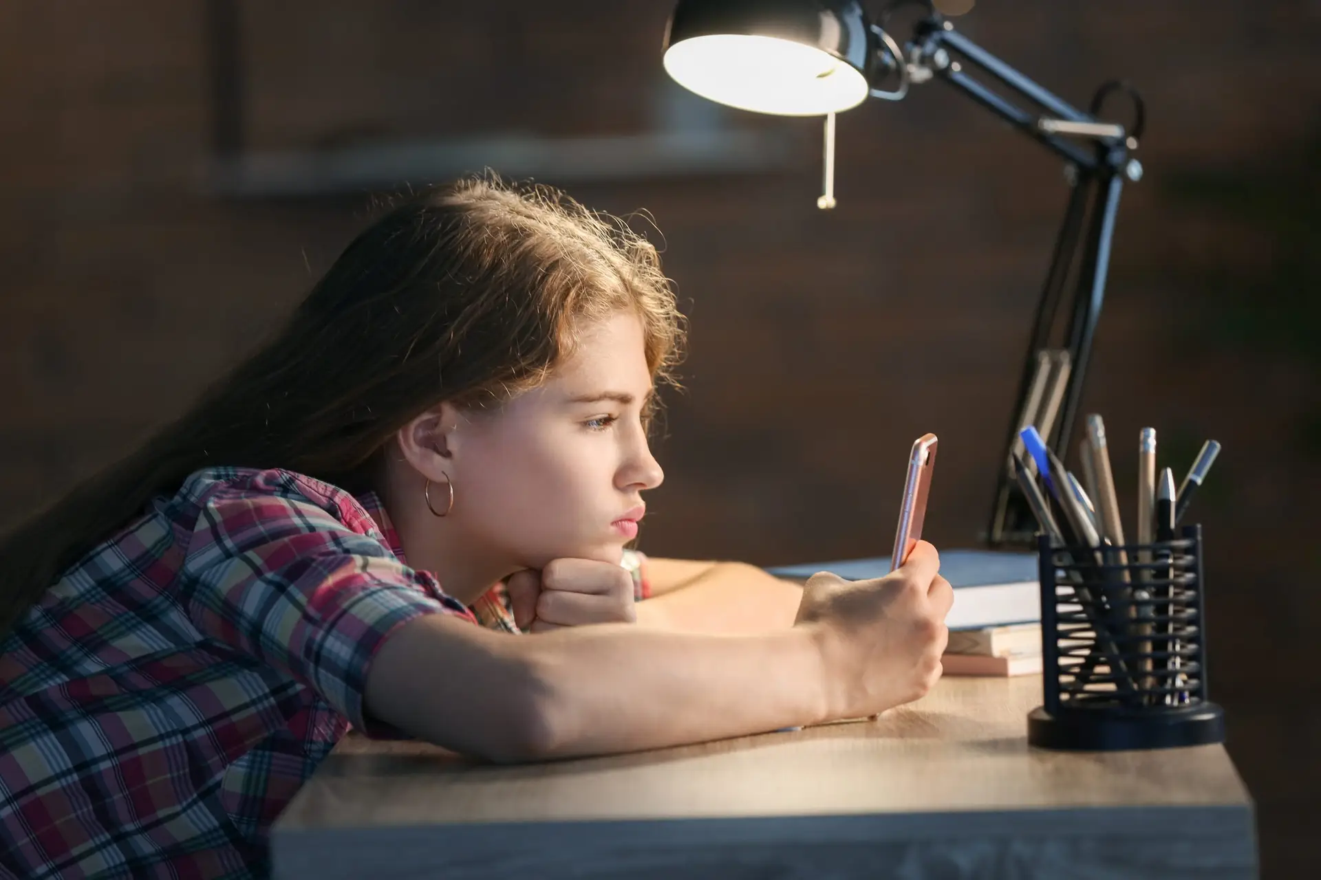 A young person, seemingly absorbed by the smartphone in their hand, sits at a desk lit by a lamp. Clad in a plaid shirt, they lean on their arm amidst pens and a blurred backdrop—an image reflecting how children can become so easily addicted to technology.