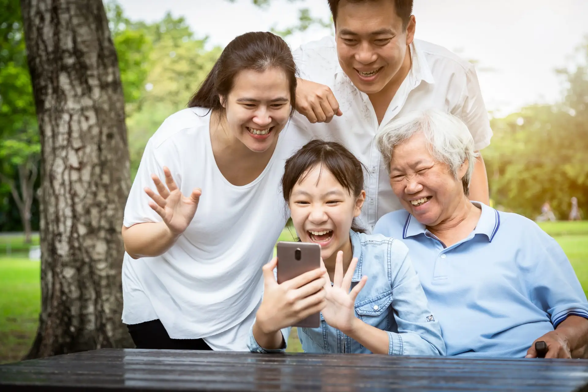 A joyful family consisting of a young child, two adults, and an elderly person gather around a smartphone, smiling and waving at the screen. They are outdoors in a park with green trees, excitedly connecting online amidst the serene natural backdrop.