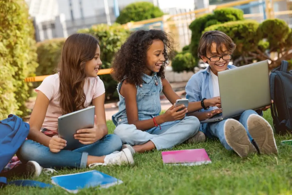 Three children sit on grass in a park, smiling as they explore the benefits of technology for kids. One holds a tablet, another a smartphone, and the third uses a laptop. Notebooks and backpacks are nearby, with trees and buildings visible in the background.