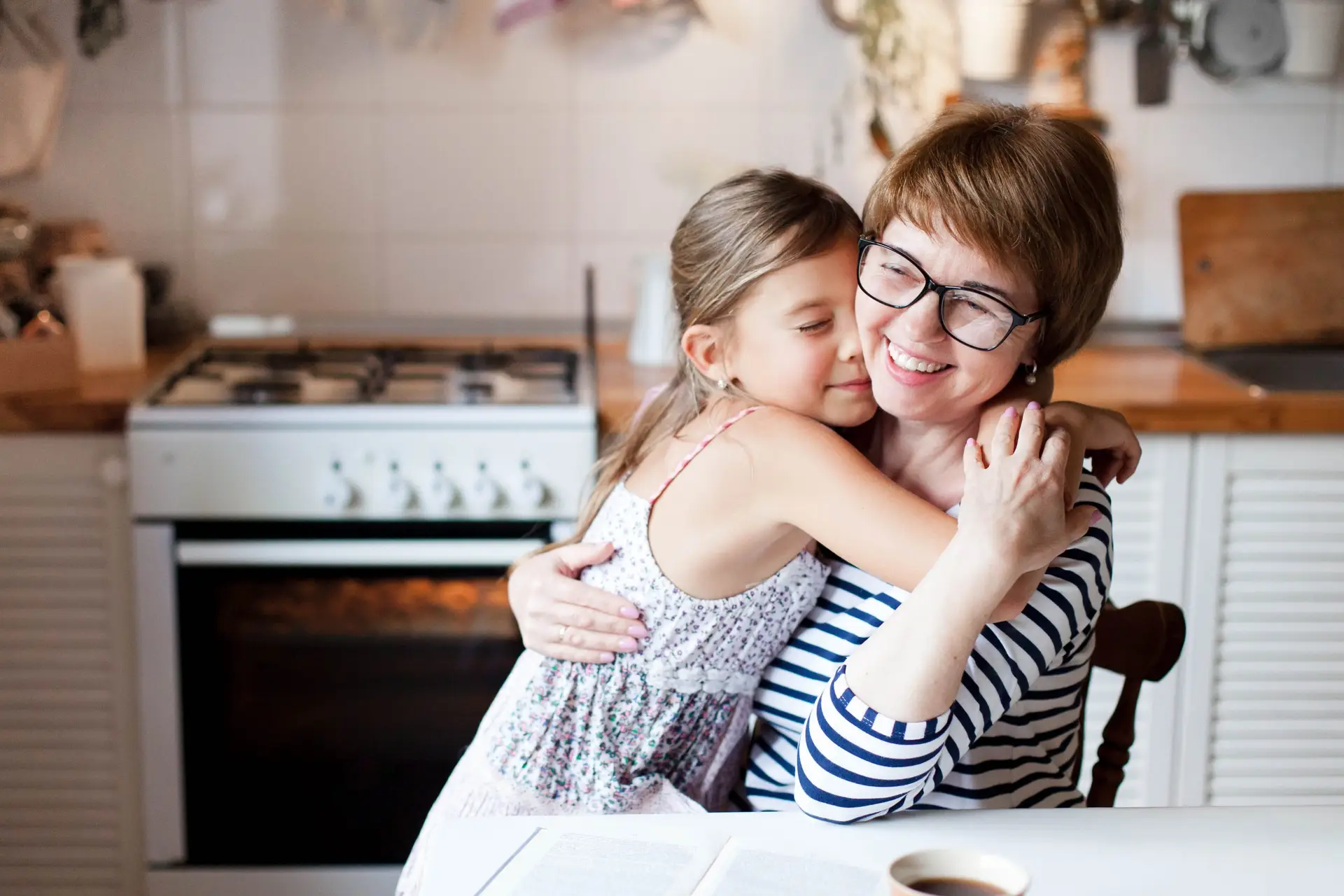 A smiling woman wearing glasses sits at a kitchen table, hugging a young girl. The scene, with its cozy backdrop of a stove and a book titled "How to Teach a Child Humility," alongside a cup, radiates warmth and affection.