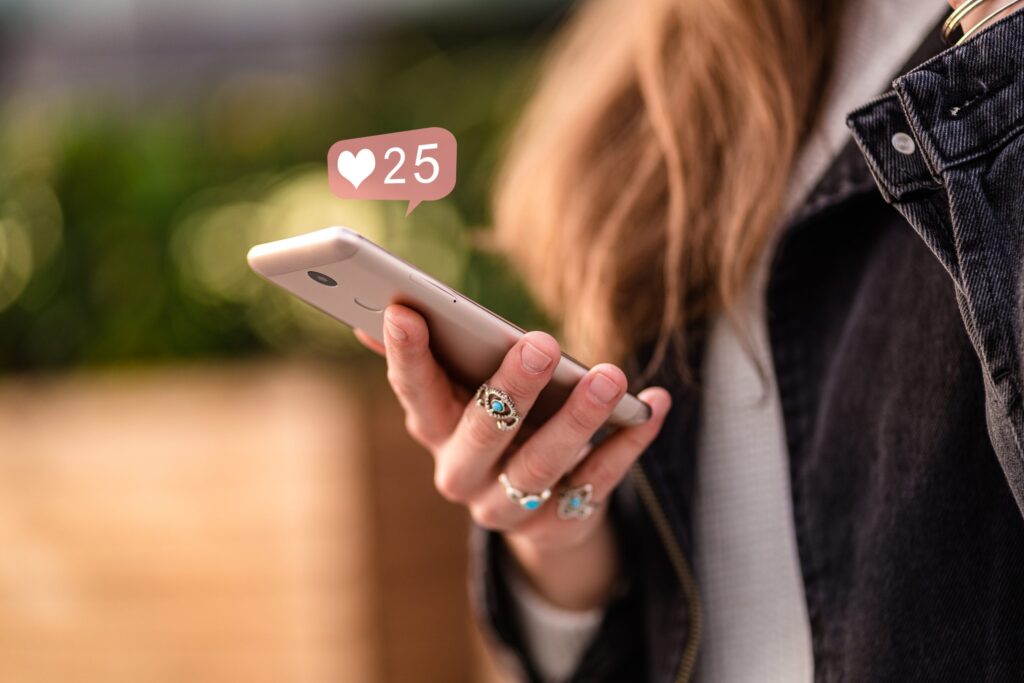 A person with long hair, wearing rings and a dark jacket, holds a smartphone. A notification with a heart icon and 25 hovers above, highlighting the social media influencer's effect on society amid blurred greenery.