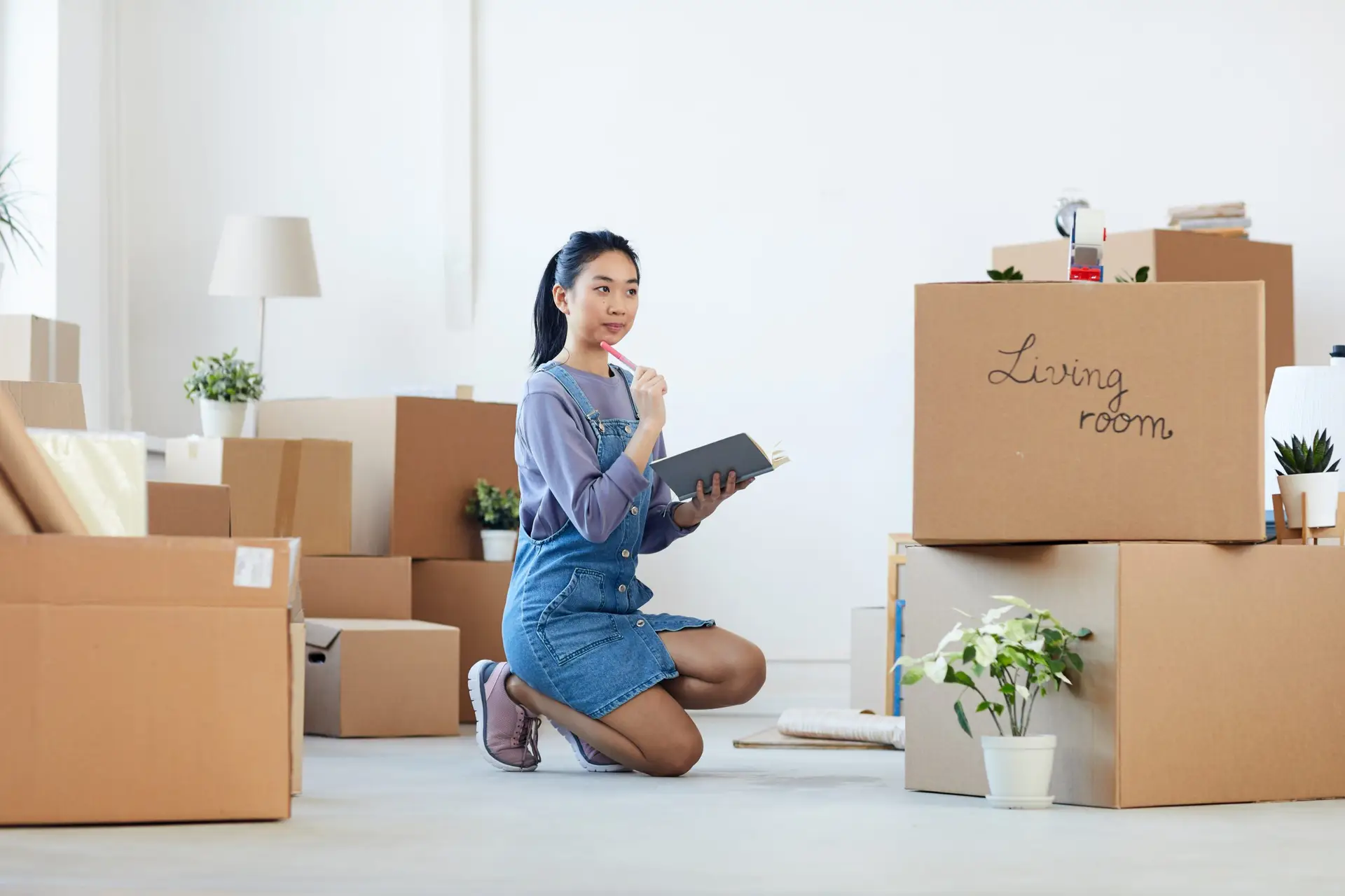 A woman kneels on the floor in a room filled with cardboard boxes, some labeled Living Room. Holding a book and pencil, she appears thoughtful and organized—perhaps pondering how to teach organizational skills to children. The room has potted plants and minimalist decor, creating a serene atmosphere.