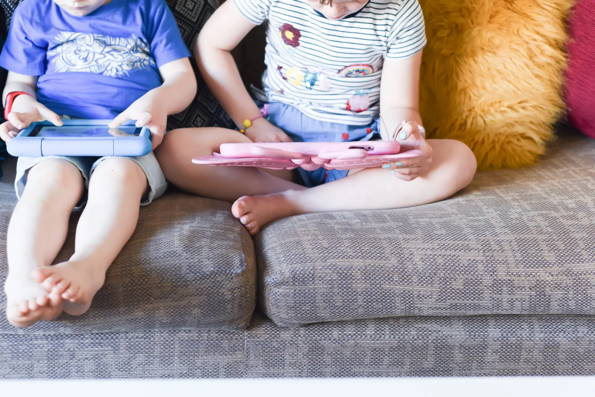 Two children sit on a couch, engrossed in free educational apps for kids on their tablets. One wears a blue shirt and shorts, while the other sports a striped shirt and skirt. Bright cushions add a cheerful backdrop to this cozy learning scene.