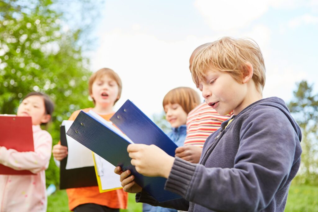 Four children are outdoors in the middle of a thrilling scavenger hunt for kids, each holding a brightly colored binder or clipboard. The sunny scene is alive with green trees, as the engaged children focus intently on their activities.