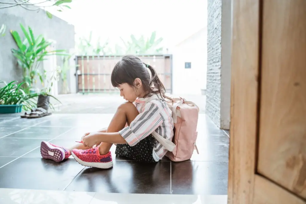 A young girl sits on the floor tying her shoelaces, perhaps unaware of the dangers of a digital footprint. She wears a striped shirt, black polka-dot shorts, and a pink backpack. Her pink shoes match the backpack. The setting is a tiled floor near a wooden door with greenery in the background.