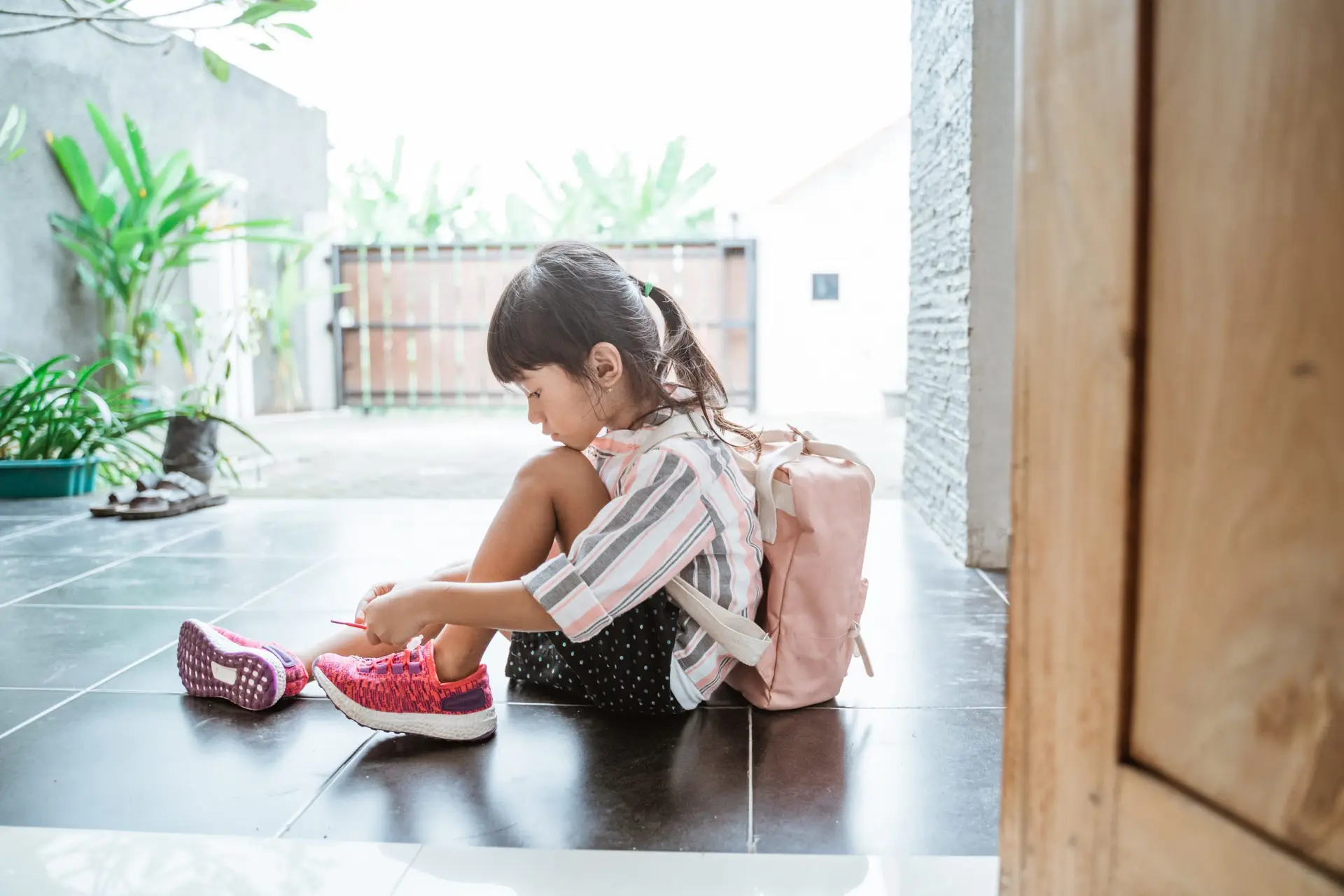 A young girl sits on the floor tying her shoelaces, perhaps unaware of the dangers of a digital footprint. She wears a striped shirt, black polka-dot shorts, and a pink backpack. Her pink shoes match the backpack. The setting is a tiled floor near a wooden door with greenery in the background.