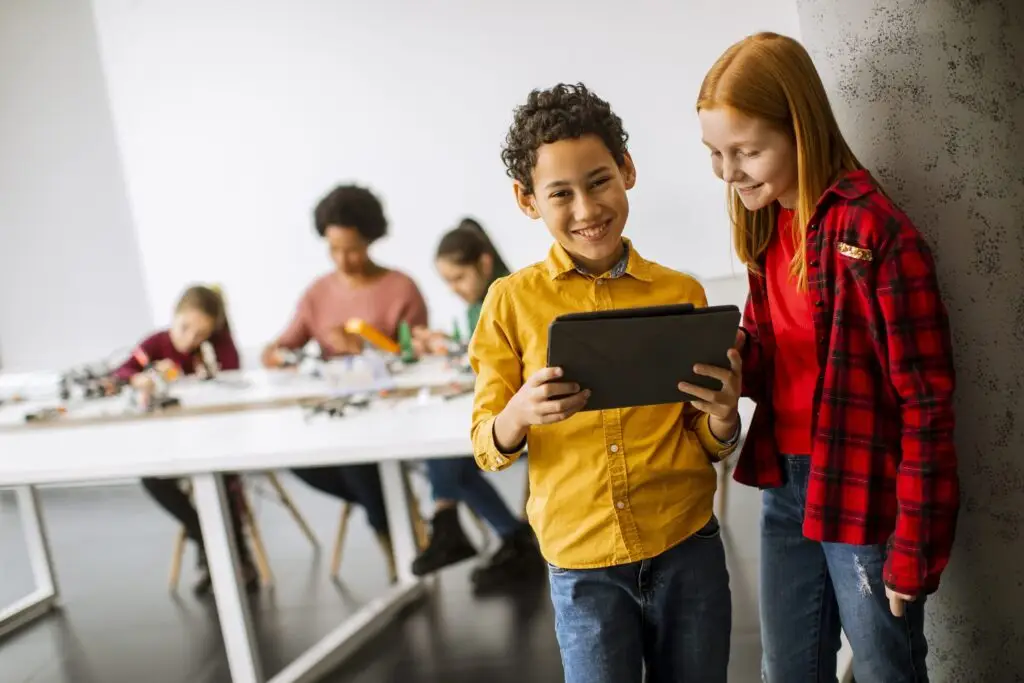 A boy in a yellow shirt and a girl in a red plaid shirt are smiling as they look at a tablet, discovering ways to do the most good they can do. Behind them, three children are seated at tables, engaged in activities in the bright room with white walls.