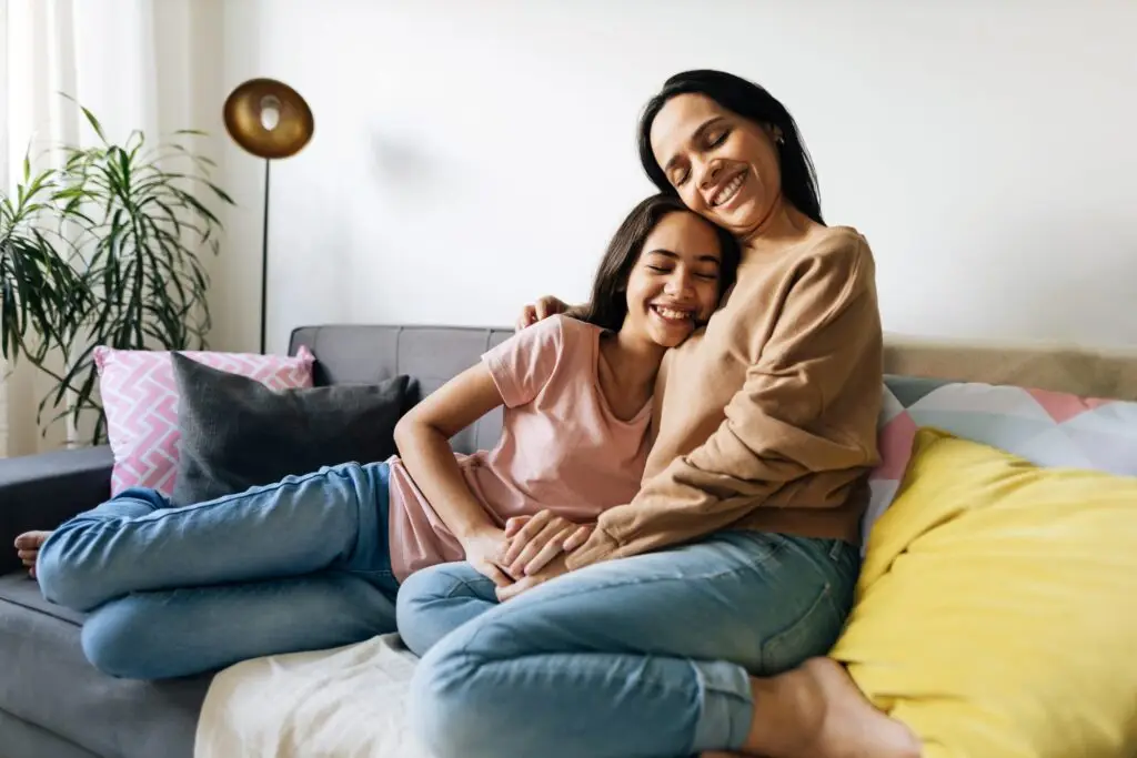 A woman and a young girl sit on a sofa, smiling and embracing each other, developing trust. The room is bright with a modern lamp and plants in the background, while colorful pillows accent the seating area.