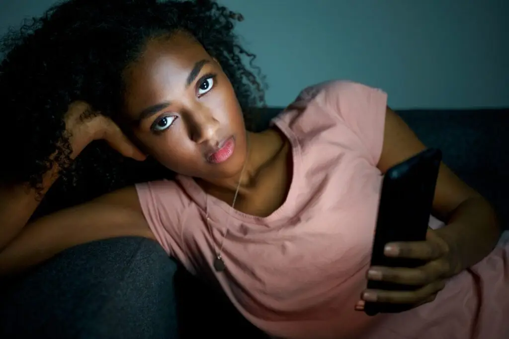 A woman with curly hair and a serious expression reclines on a sofa, holding a smartphone. She wears a pink shirt and gazes intently at the camera, embodying an aura of perfectionism in the dimly lit room.